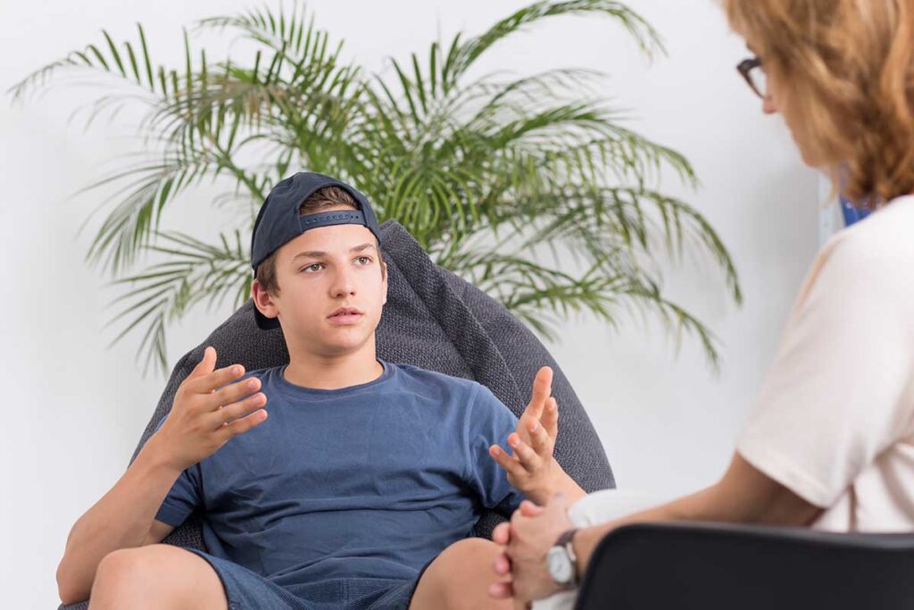a teen boy sits while talking to his therapist asking her about the benefits of cognitive-behavioral therapy